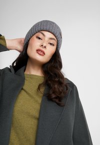 Bonnet en tricot gris, texture côtelée, porté avec un manteau gris foncé et un pull vert olive. Tissu doux, design décontracté, marque discrète visible.