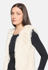 Kamizelka ze sztucznego futra w kolorze kremowym o puszystej fakturze, z otwartym przodem i bez rękawów, noszona na czarnej bluzce z długim rękawem. Gładkie włosy, neutralny wyraz twarzy.