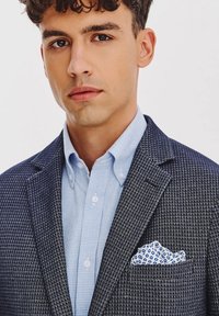 Granatowy marynarka w kratę houndstooth z wyciętymi klapami, w połączeniu z jasnoniebieską koszulą w kratę i białą poszetką z niebieskimi wzorami kwiatowymi.