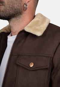Uomo che indossa una giacca marrone con colletto in pelliccia sintetica beige, camicia bianca sotto e orecchino a cerchio argento all'orecchio sinistro.