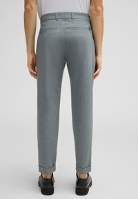 Pantaloni grigi su misura con una finitura textured, orli con risvolto e due tasche posteriori. Indossati con scarpe nere, dal design minimalista senza logo visibile.
