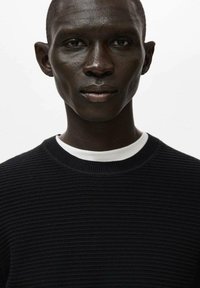 Maglione nero in maglia costina con scollo rotondo, sovrapposto a una maglietta bianca con scollo a girocollo. Il volto del modello presenta pelle liscia e zigomi definiti.