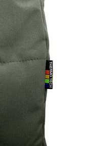 Grijs stoffen product met een gequiltde textuur. Voorzien van een zwart label met "THERMO-TECH" en een regenboogstreepontwerp. Eenvoudige rechthoekige vorm.