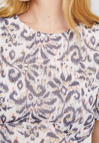 Halenka s květinovým vzorem, která má bílý základ s pastelově fialovými a broskvovými motivy, texturovaný povrch a zakulacený výstřih.