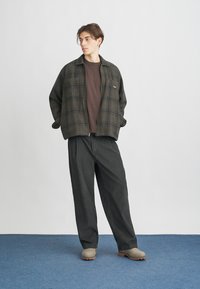 Károvaná zipová bunda v tmavozelenej a hnedej farbe, skombinovaná s hnedou košeľou a čiernymi širokými nohavicami. Béžové topánky s gumovou podrážkou s textúrou.