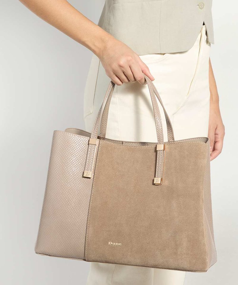 Femme tenant un grand sac cabas beige avec des côtés texturés et un panneau central en daim, portant un pantalon clair et un gilet sans manches.