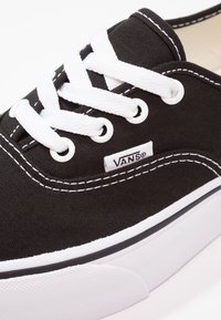 Svart canvas sneakers med vita snörningar, med en Vans-logotyp och kontrasterande vita sömmar. Sålen är vit med en strukturerad finish.