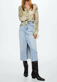 Blusa verde a fantasia con maniche lunghe, abbinata a una gonna midi in denim blu chiaro con uno spacco frontale. Stivali neri con tacco completano il look.