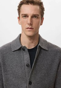 Szary prążkowany kardigan z kołnierzykiem, z przodu zapinany na guziki i o miękkiej fakturze, noszony na czarnej koszuli.