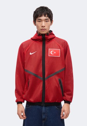 Młody mężczyzna w czerwonej kurtce Nike z naszywką tureckiej flagi i czarnymi detalami na zamkach, stoi na jednolitym jasnym tle.