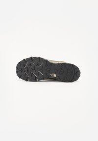 Trail shoe sole with textured black rubber, featuring a rugged tread pattern for traction and reinforced logo detail in neutral tones.