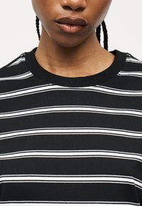 Κοντινό πλάνο ενός ατόμου με σκούρα πλεξούδα που φοράει πουκάμισο με οριζόντιες μαύρες και άσπρες ρίγες.