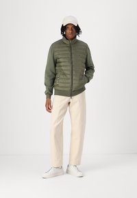 Veste verte rembourrée avec fermeture éclair à l'avant, manches et col côtelés, associée à un pantalon beige et des baskets blanches, sur un fond blanc.
