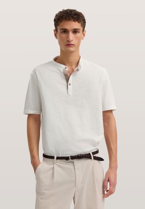 Junger Mann trägt ein weißes Henley-Shirt mit kurzen Ärmeln, das in beige Hose gesteckt ist, mit geflochtenem dunkelbraunem Gürtel, Hände in den Taschen.