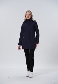 Marineblauwe waterdichte jas met een hoge kraag, verstelbare koordjes en zijzakken; gecombineerd met zwarte broeken en witte sneakers.