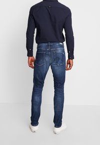 Tmavě modré džíny z denimu s úzkým střihem, vybavené dvěma zadními kapsami a značkovou nášivkou na pase, doplněné námořnicky modrou košilí.