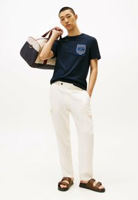 T-shirt blu navy con grafica del logo, pantaloni cargo beige e sandali marroni con cinturino; trasportando una borsa bicolore con dettagli marroni.