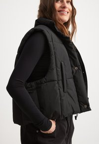 Must must vest kõrge kraega, tikandiga disain, ja küljetaskudega. Kantud pika varrukaga pluusi peal, millel on tõmblukk ja nupud.