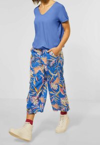 Top blu V con scollo a V abbinato a pantaloni blu larghi con un motivo tropicale colorato e tasche laterali. Indossato con sneakers beige alte.