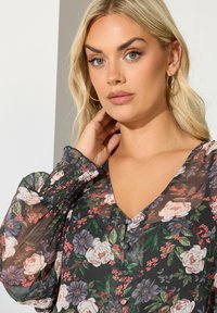 Donna bionda con occhi azzurri chiari, che indossa orecchini a cerchio dorati e una blusa trasparente nera a fiori con maniche lunghe a sbuffo e bottone frontale.