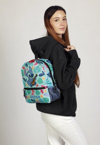 Mochila de tela azul clara, con un diseño tropical que presenta patrones de hojas y flores, así como un personaje llamado Stitch.