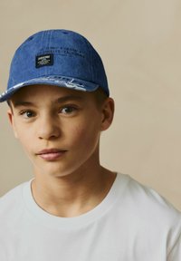 Blå denimkasket med grafiske detaljer og et sort mærke med koordinater og tekst, båret med en simpel hvid T-shirt.