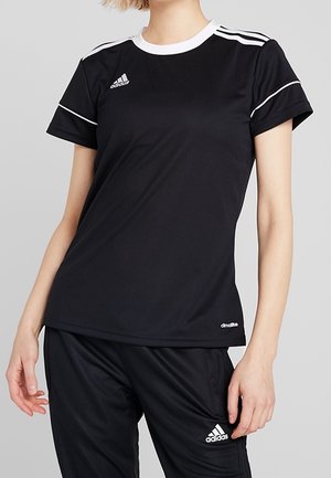 Person iført sort Adidas sportstøj med hvide striber på ærmer og bukser, stående mod en ensfarvet grå baggrund.