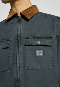 Mand iført en mørkeblå zip-up jakke med brunt kordfløjlskrag og to brystlommer, hvoraf den ene har Lee Workwear-logoet.