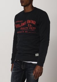 Felpa nera con testo grafico rosso, colletto a girocollo e polsini a coste, abbinata a jeans in denim blu. Presenta un tessuto testurizzato e un'etichetta con logo.