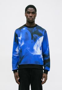 Hombre negro vestido con un sudadera azul y negra con patrones abstractos en blanco, de pie frente a un fondo blanco liso.