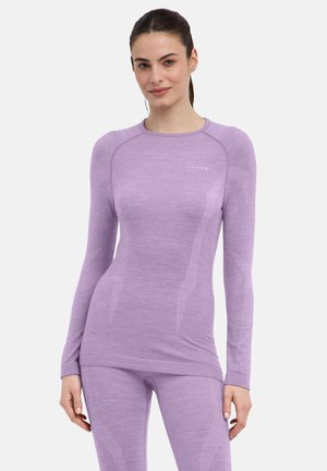 Vrouw met haar naar achter gebonden, die een aansluitend lavendelkleuren top met lange mouwen en bijpassende leggings draagt tegen een effen achtergrond.