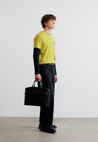 Jeune homme portant des chemises à manches longues superposées jaunes et noires, un pantalon noir et des chaussures, tenant une mallette noire, debout sur un sol en béton.