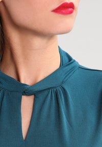 Turkusowa bluzka z węzłem na dekolcie, wykonana z miękkiego, drapowanego materiału z detalem w kształcie łezki. Gładka tekstura z delikatnym połyskiem.