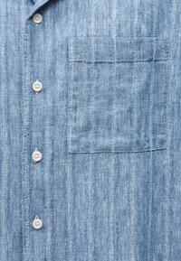 Υφασμάτινο πουκάμισο ανοιχτό μπλε με υφή, κάθετες ρίγες, λευκά κουμπιά και τετράγωνη τσέπη στο στήθος στη δεξιά πλευρά.