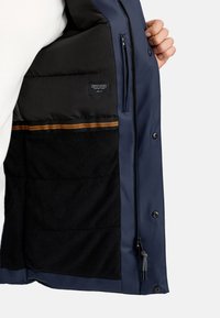 Marineblauwe jas met een gevoerde binnenkant, zwarte fleece voering, ritszak en een bruine gestreepte accent langs de voering.