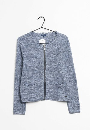 Cardigan en tricot bleu-gris à manches longues, avec une fermeture éclair à l'avant, deux poches avant aux bords effilochés, suspendu à un cintre en fil métallique noir.