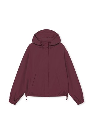 Jonge man met een bordeauxrode capuchonjas met elastische boorden en een zwarte broek, staand tegen een effen witte achtergrond.