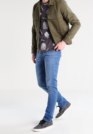 Giacca verde con bottoni, t-shirt grigia con stampa, jeans aderenti blu e scarpe da ginnastica grigie con suole bianche, con lacci e design sottile.