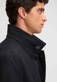 Cappotto in misto lana nero con colletto alto, caratterizzato da una texture liscia e un dettaglio con un singolo bottone nero sul colletto.