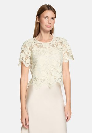 Spitzentop in Off-White mit kurzen Ärmeln, geschwungenem Saum und Blumenmuster. Getragen über einer glatten, cremefarbenen Unterlage.