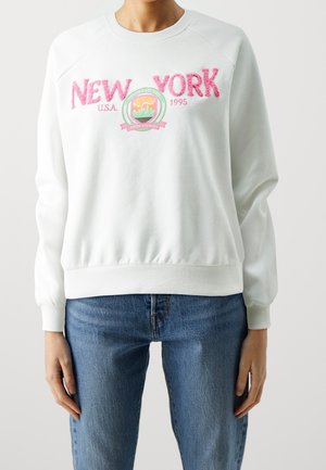 Persoon draagt een witte trui met roze tekst en embleem "New York U.S.A. 1995", gecombineerd met een blauwe spijkerbroek.