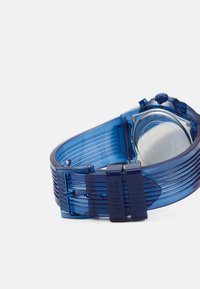 Blauwe siliconen horloge met geribbelde band, zilverkleurige onderdelen en een ronde kast. Heeft een doorzichtige achterkant, meerdere verstelgaten en blauwe knoppen.