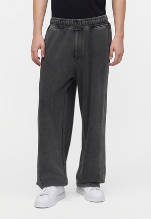 Person iført løse mørkegrå joggingbukser og hvide sneakers mod en ensfarvet lys baggrund.