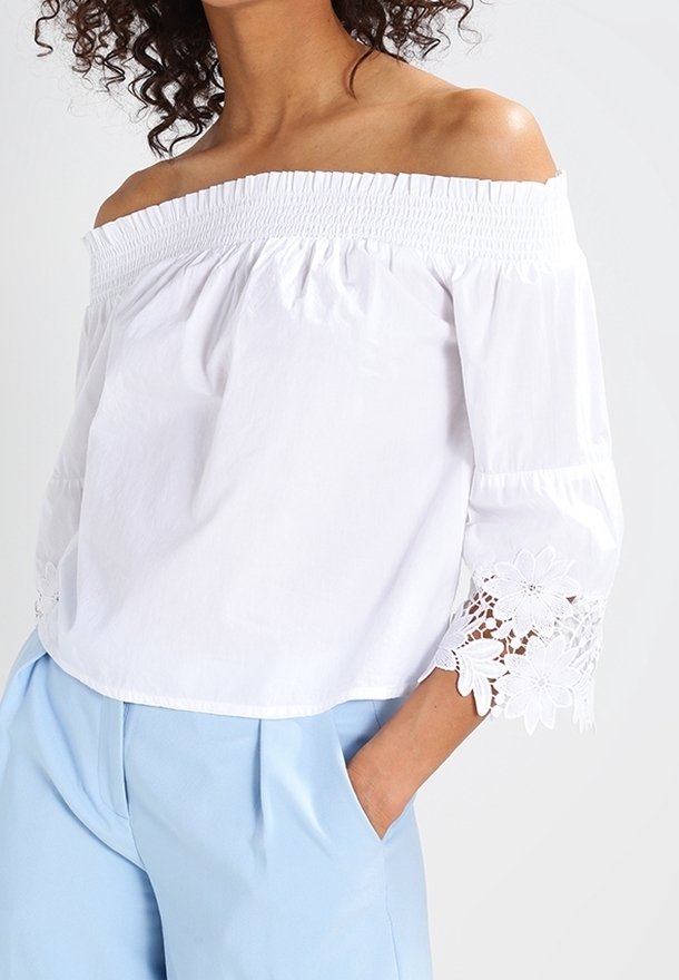 Femme portant un chemisier blanc épaules dénudées avec des poignets en dentelle florale et un pantalon bleu clair taille haute, une main dans la poche.