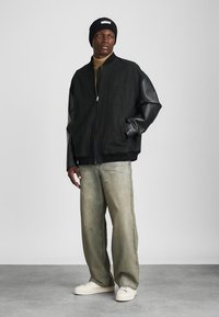 Bărbat îmbrăcat cu o jachetă bomber neagră cu mâneci din piele, pulover bej cu guler înalt, blugi gri deschis, adidași albi și o căciulă neagră, stând pe un podea gri.