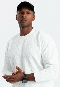Vit, texturerad sweatshirt med upphöjd bokstavsmönster, rund halsringning och långa ärmar. Svart klocka med rund urtavla på handleden.