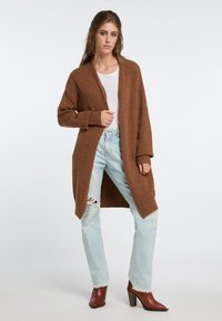 Rudos megztinis su laisvu siluetu, atviru priekiu ir ilgomis rankovėmis, derinamas su šviesiai mėlynomis nusidėvėjusiomis džinsais ir rudomis aukšto kulno auliniais batais.