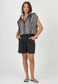 Leopartmustercropped Weste mit Kapuze, schwarzen Trägern und einer glatten Textur, kombiniert mit schwarzen, locker sitzenden Shorts und Flip-Flops.