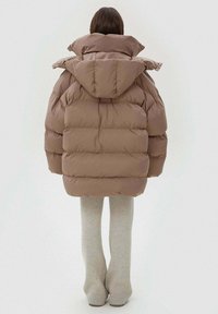 Brązowa puchowa kurtka z wysokim kołnierzem i kapturem, z pikowanym wzorem i grubym wypełnieniem. W połączeniu z jasnoszarymi dzianinowymi legginsami.