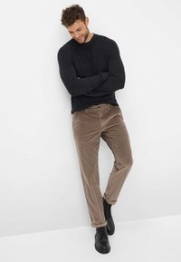 Bruine ribfluwelen broek met een rechte snit en opgerolde manchetten, gecombineerd met een donkere trui en zwarte veterschoenen, tegen een witte achtergrond.